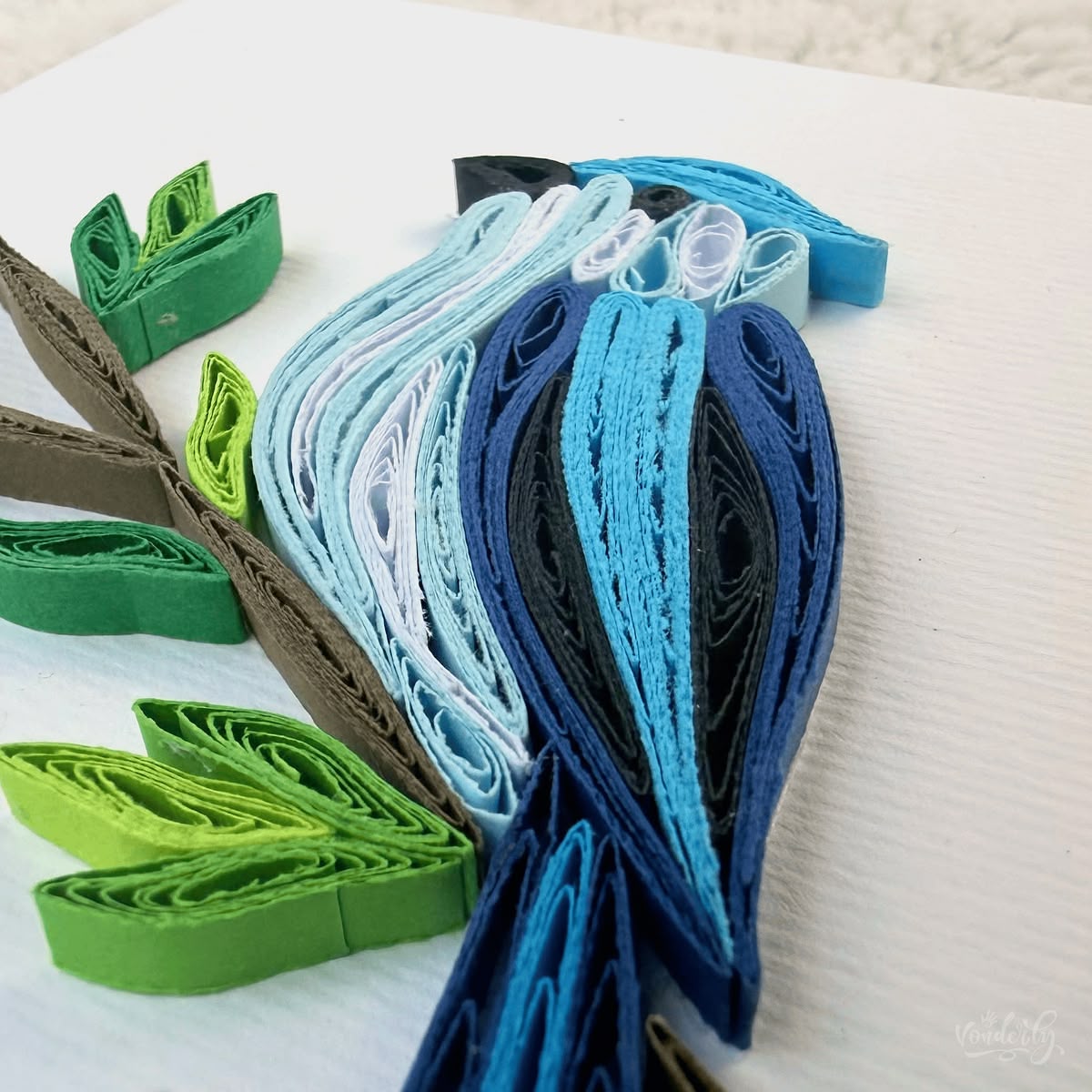 Close-up macro of paper quilling on blue jay card, three blue tones edged with black and white for plumage, plus two green tones for leaves