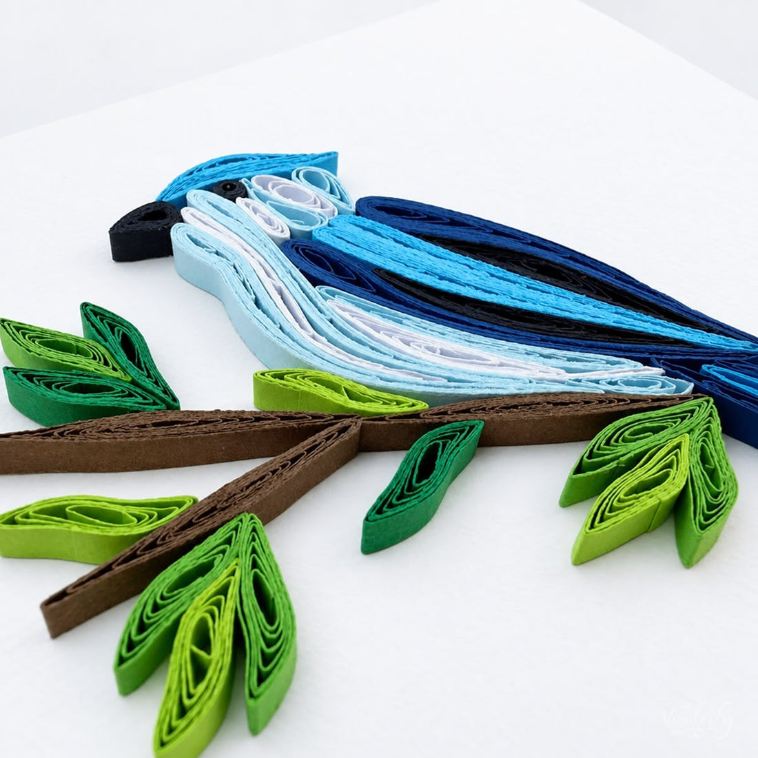 Blue Jay quilled greeting card with a crested blue songbird, white chest and black beak, on a brown branch with green leaves.