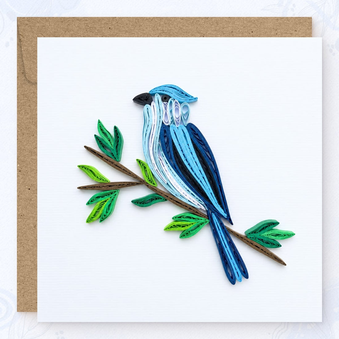 Blue Jay quilled greeting card with a crested blue songbird, white chest and black beak, on a brown branch with green leaves.