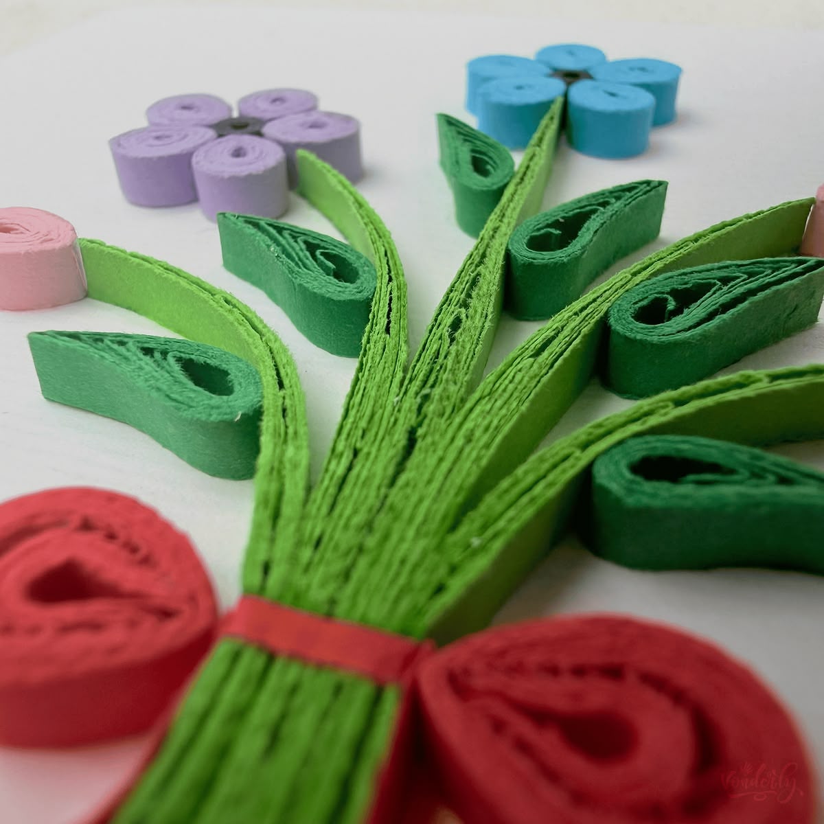 Close-up macro of paper quilling on bouquet card, gathered green stems and red ribbon tie with adjacent flower elements visible