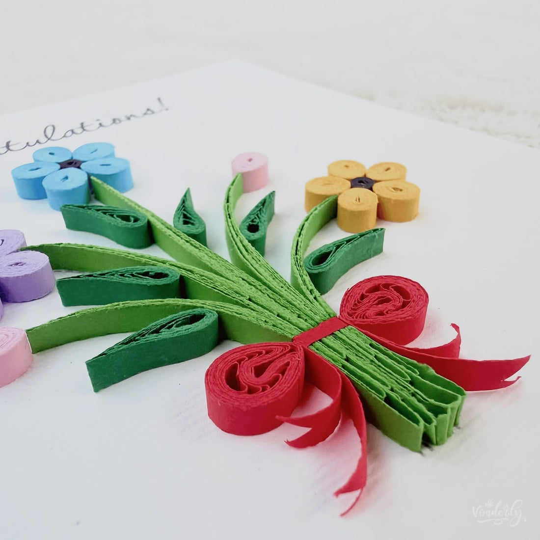 Handmade quilled congratulations greeting card with three flowers (blue, lavender, orange), two pink buds, and a red ribbon bow on gathered green stems, "Congratulations" printed in calligraphic script, paper art by Vonderly