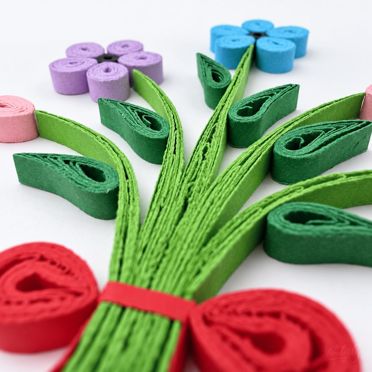Close-up of purple and blue coiled paper petals with black centers above green stems and a red quilled bow.