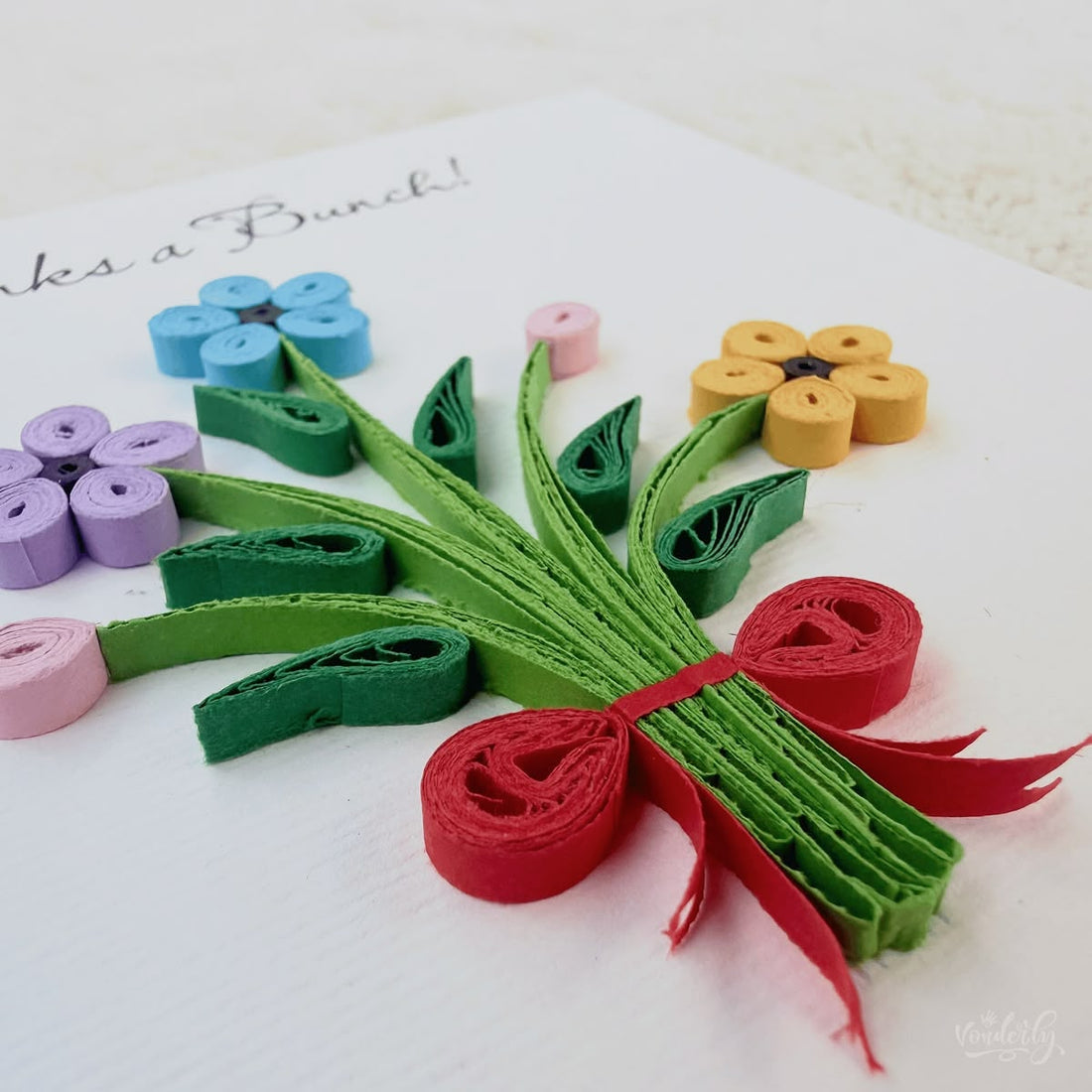 Handmade quilled thank you greeting card with three flowers (blue, lavender, orange), two pink buds, and a red ribbon bow on gathered green stems, "Thanks a Bunch" printed in calligraphic script, paper art by Vonderly