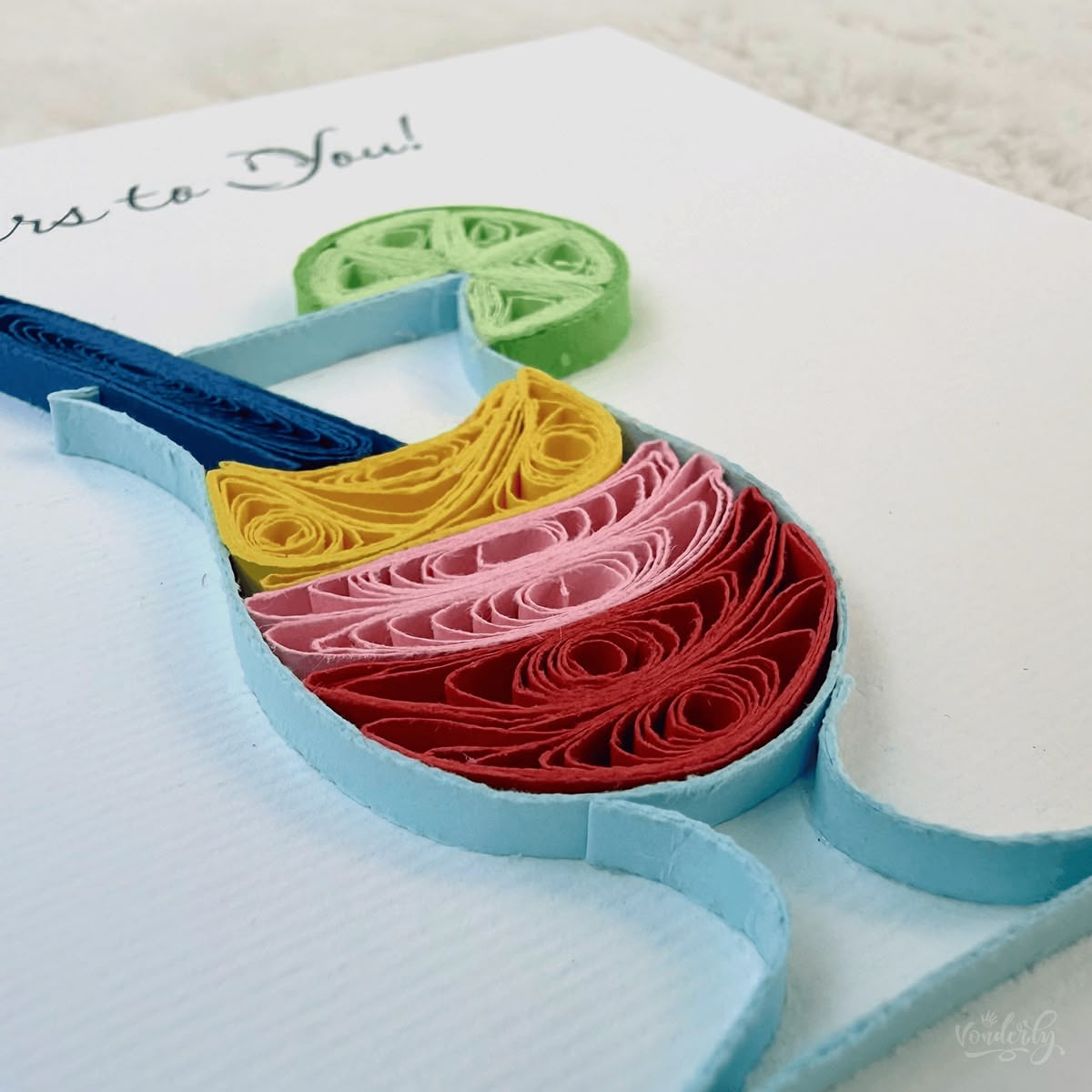 Angled view of quilled cocktail congratulations card showing 3D raised paper art, the layered drink, navy stir stick, and green lime garnish standing off the card surface