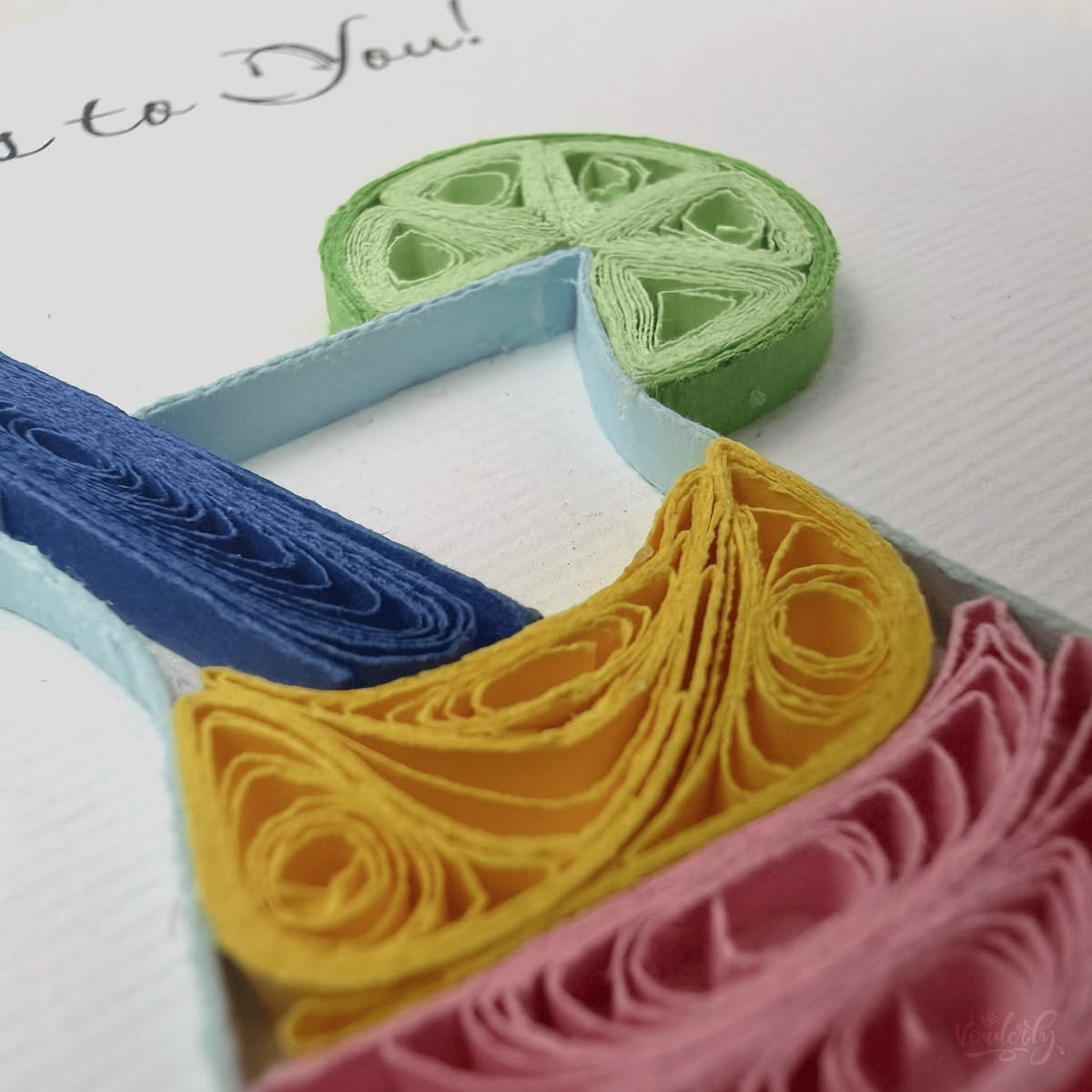 Close-up macro of paper quilling on the cocktail card, scrollwork fill in red, pink, and gold drink layers, navy swizzle stick, and green husked lime wheel