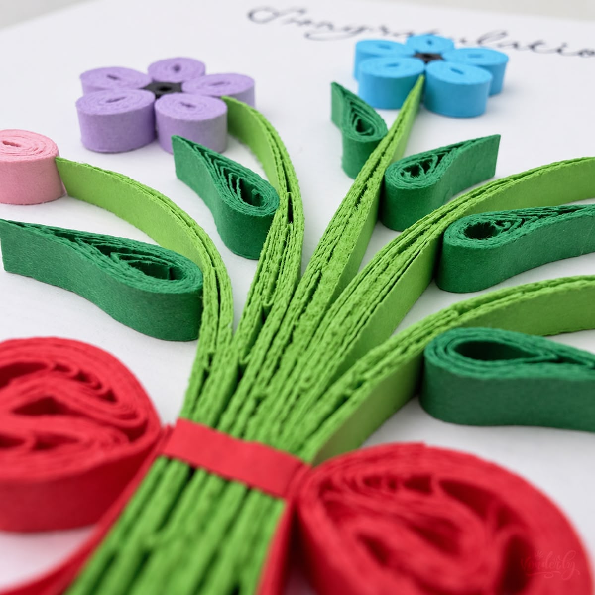 Close-up of green paper leaves and a red bow beneath purple and blue quilled flowers.