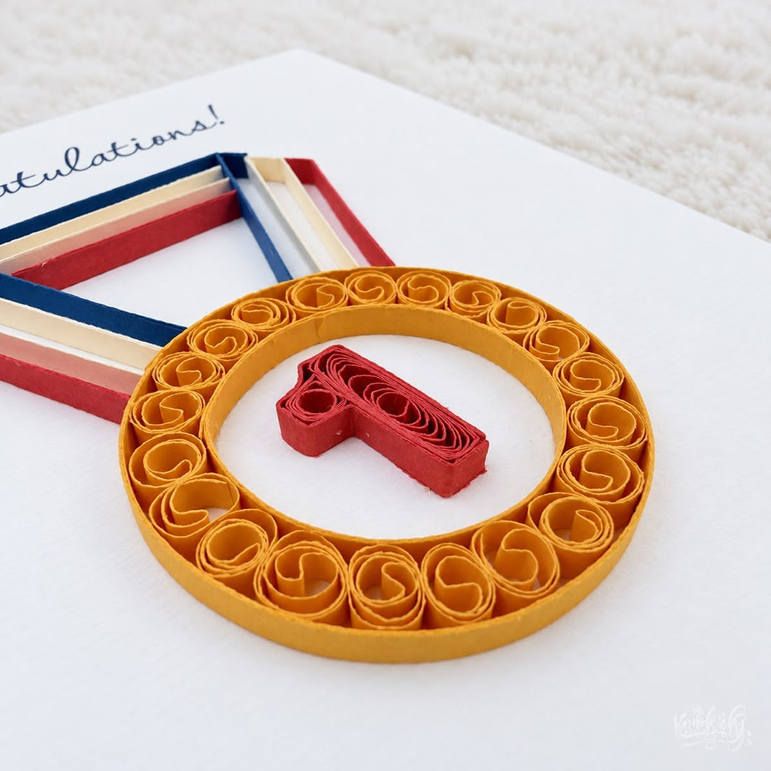 Quilled congratulations card with a gold paper medal and red number 1 on a navy, cream, and red ribbon.
