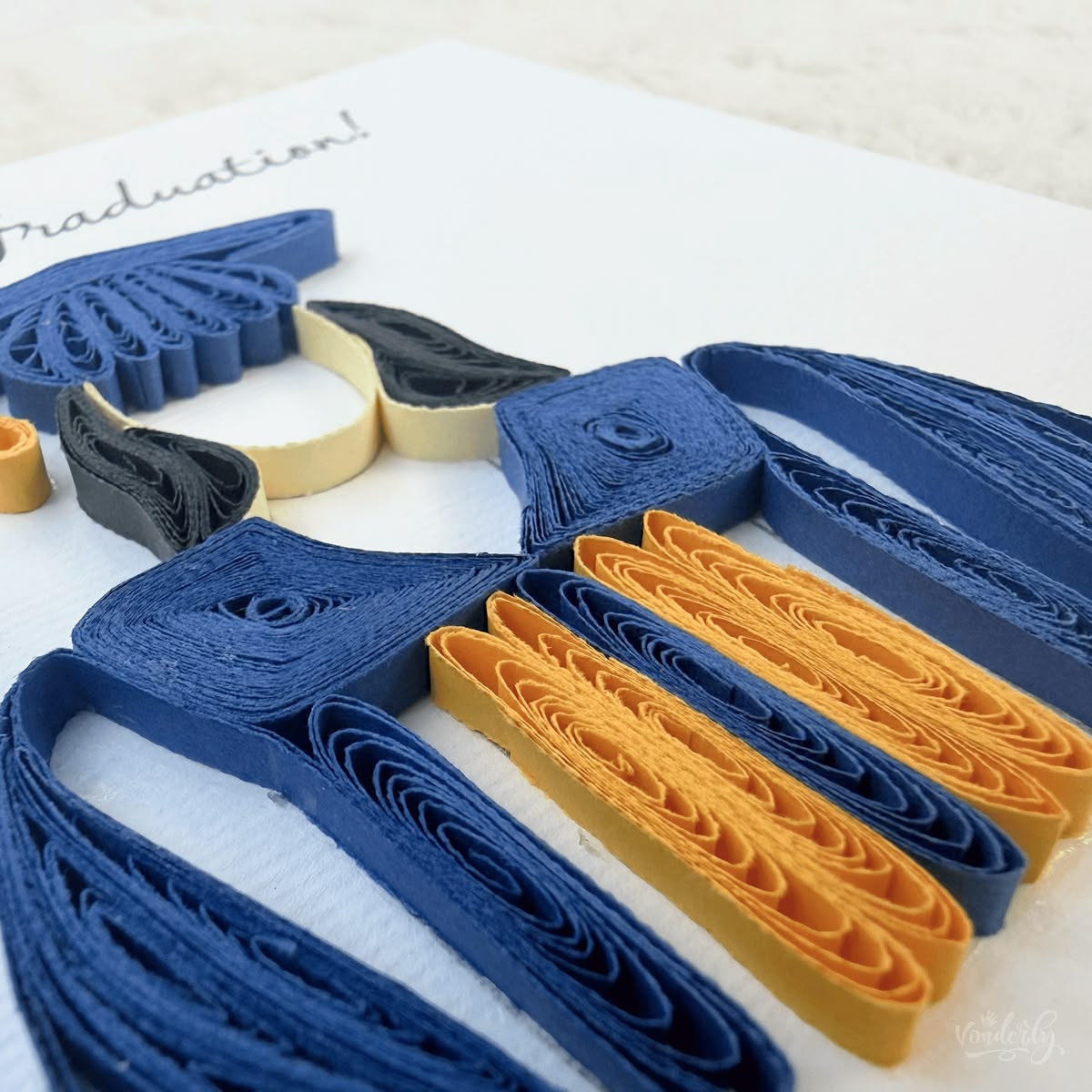 Angled view of quilled graduation card showing 3D raised paper art, cap dome and gold stole detail visible