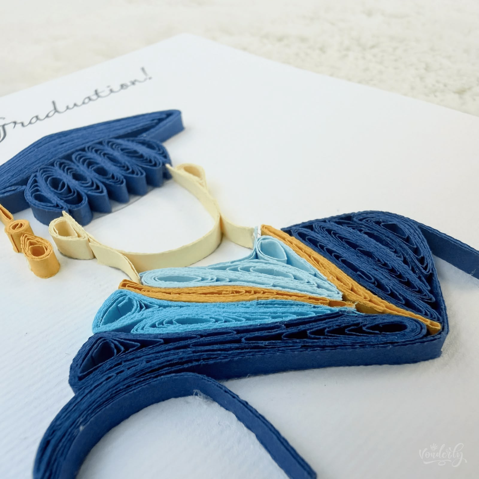 Angled view of quilled graduation card for him showing 3D raised paper art, navy gown and light blue hood detail