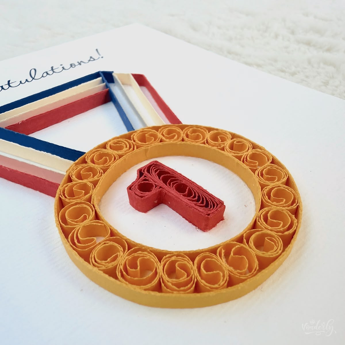 Angled view of quilled congratulations card showing 3D raised paper art, gold medal ring with red number 1 and layered ribbon visible