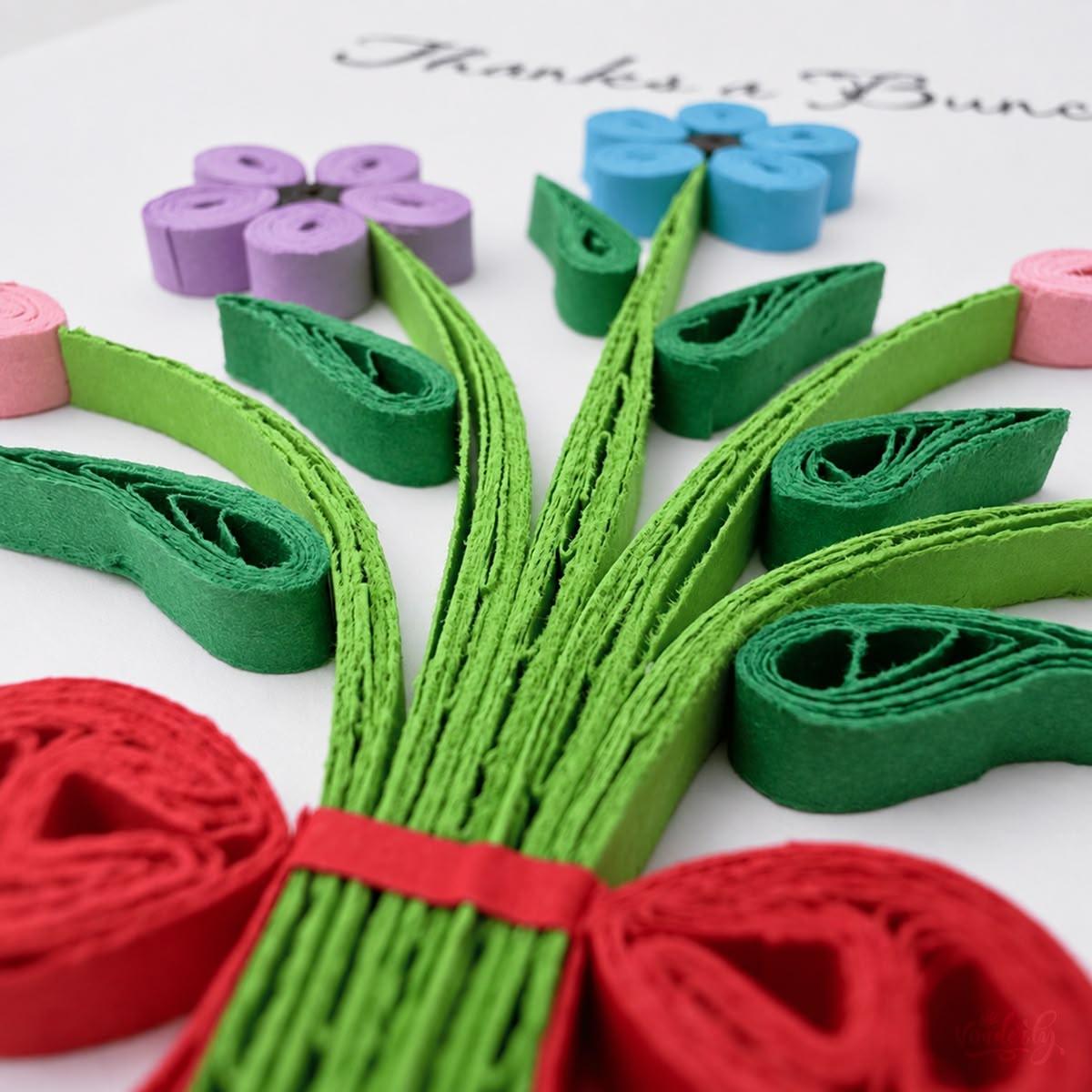 Close-up of green paper leaves and a red bow beneath purple and blue quilled flowers.
