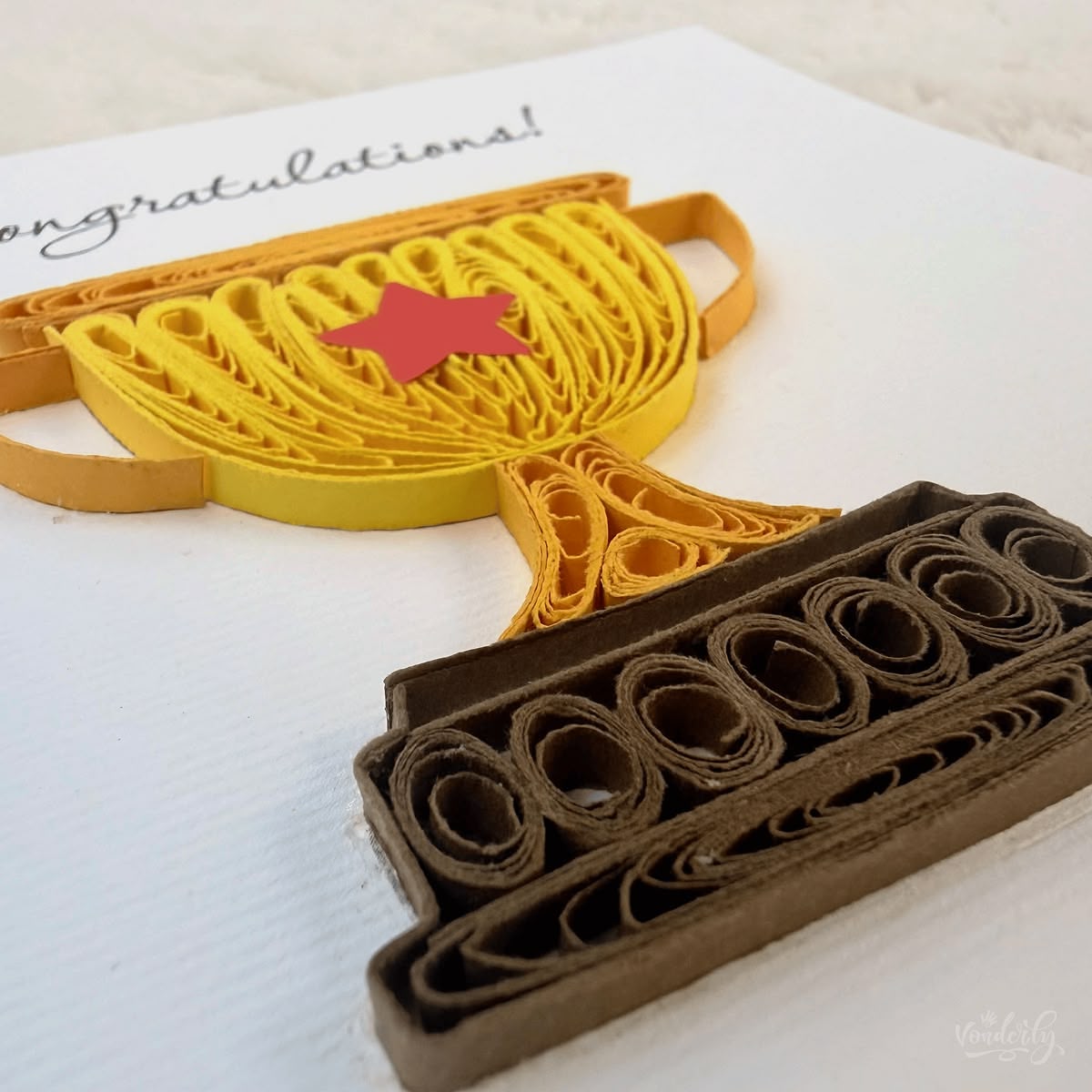 Angled view of quilled congratulations trophy card showing 3D raised paper art, layered cup body with handles and packed-coil base visible
