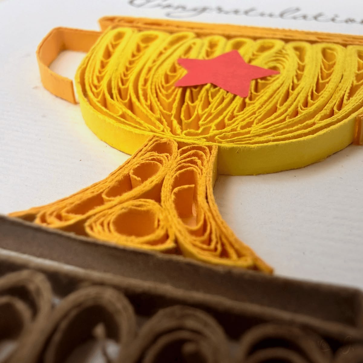 Close-up macro of paper quilling on trophy card, curved yellow paper loops forming the cup body and packed dark coils forming the rectangular base