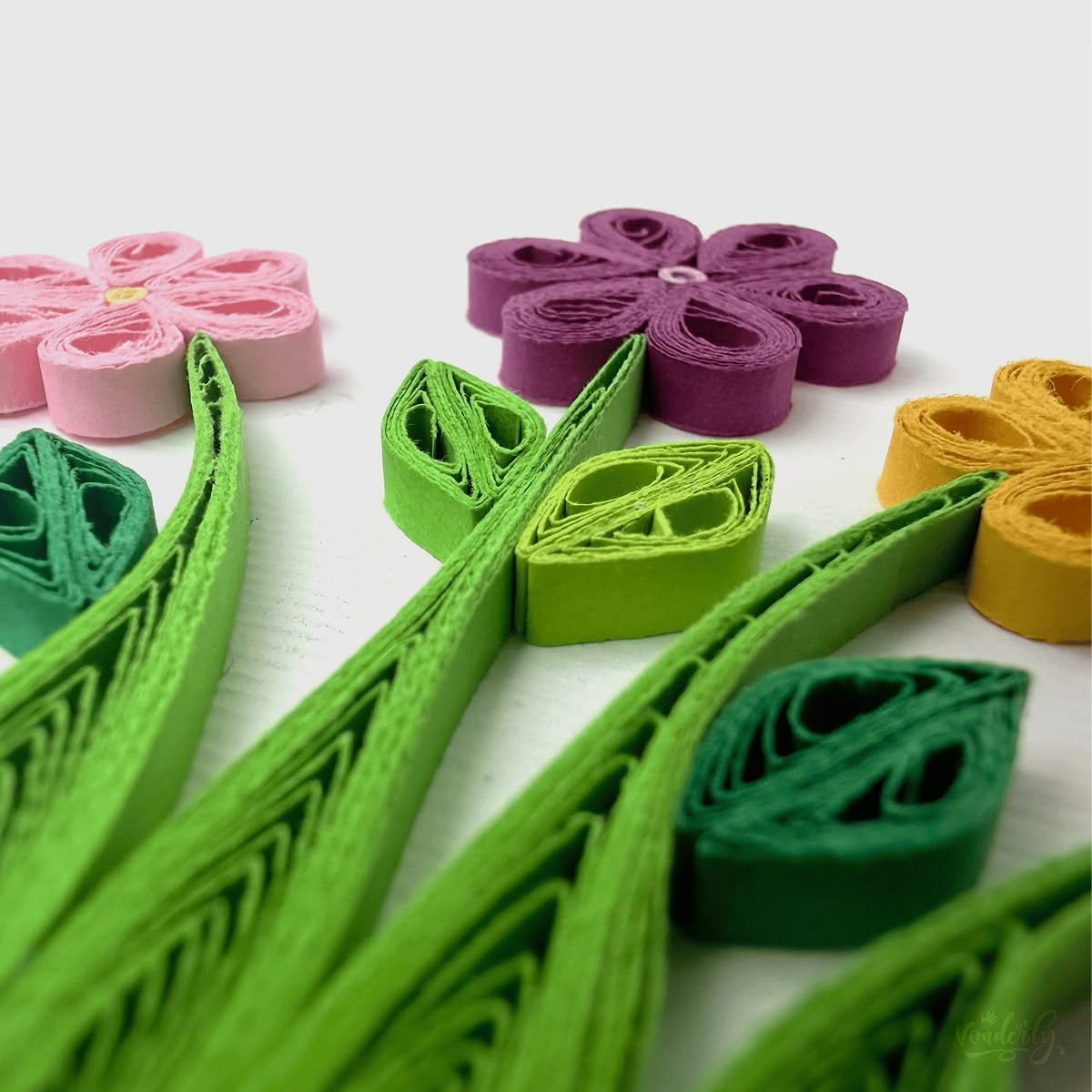 Close-up macro of paper quilling on wildflowers card, six-petal flowers in pink, purple, and yellow with stems and leaves in three greens